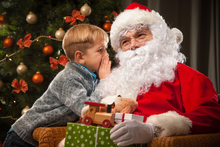 A child whispers into Santa's ear. Santa holds a pile of presents on their lap.