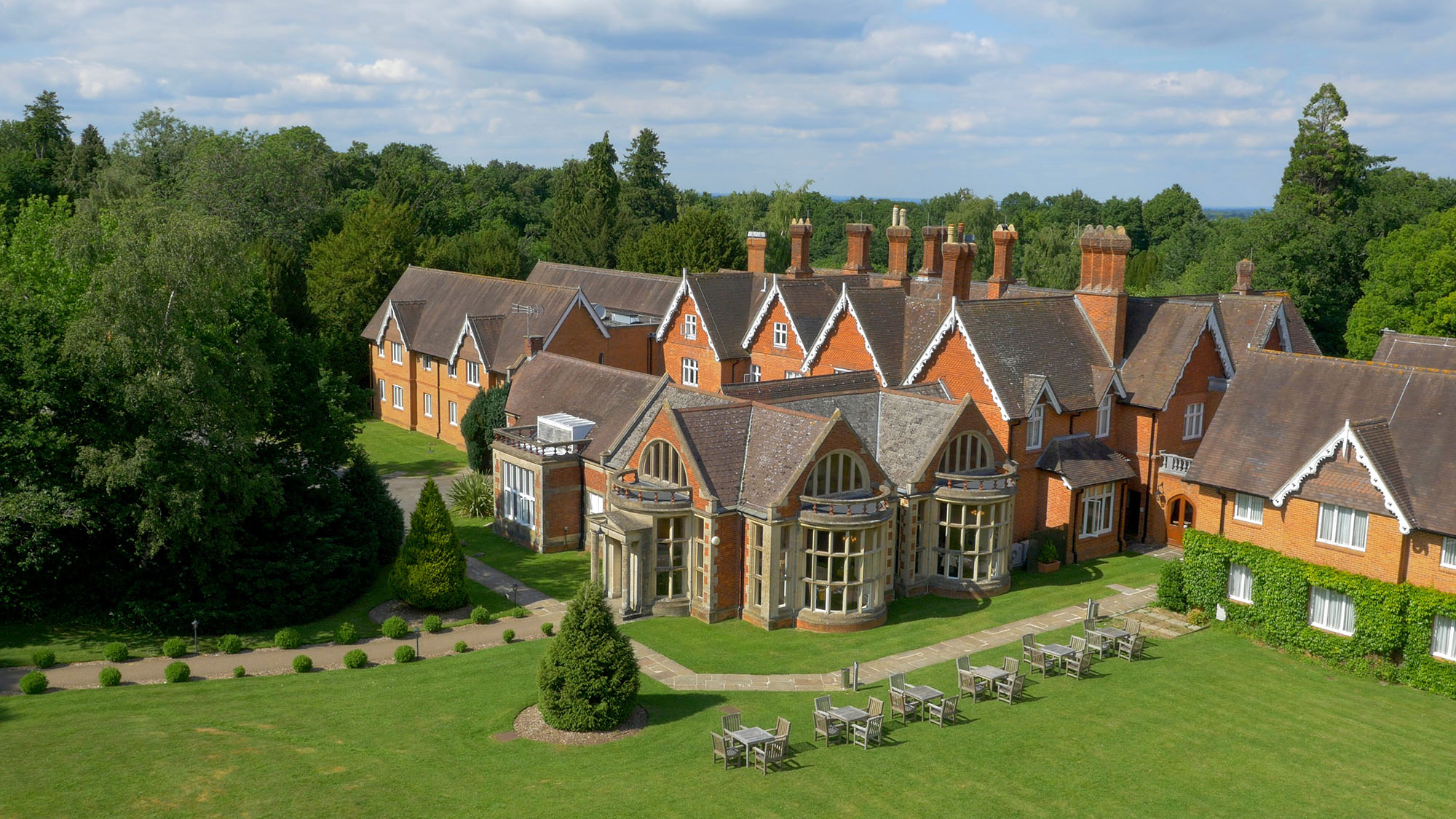 A red brick building with brown roof. It is surrounded by large mature trees and lawn. There are tables and chairs out on the lawn.
