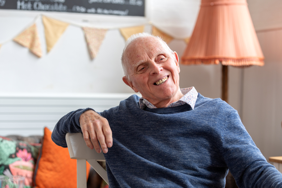 A man sits relaxed on a chair, with one arm slung over the back and is smiling looking to the side.