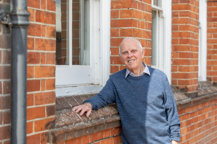A main stands outside with his arm resting on the wall and he looks at the camera.