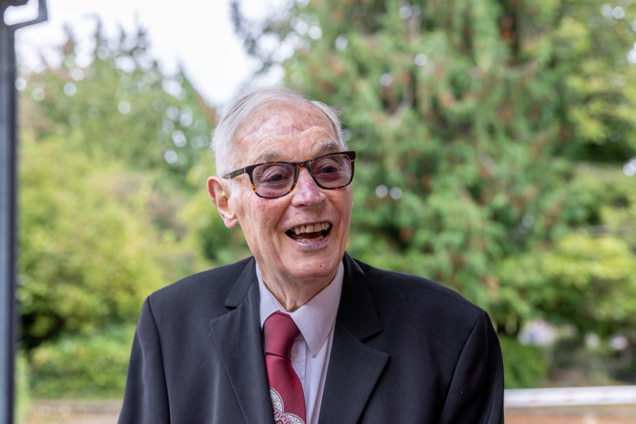 A male smiles and looks away from the camera. In the background are green trees.