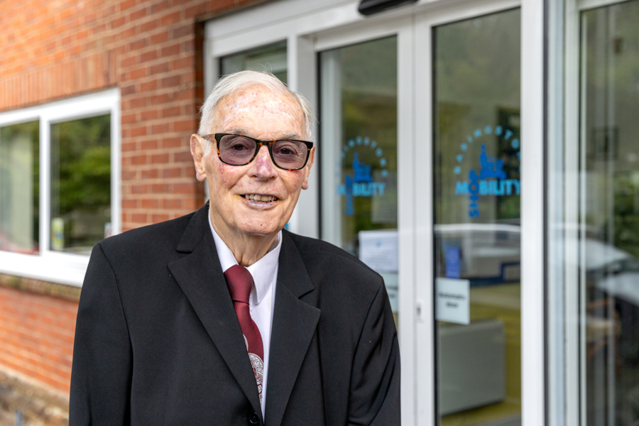 A man stands in a suit with a red tie. He's outside the shopmobility building.