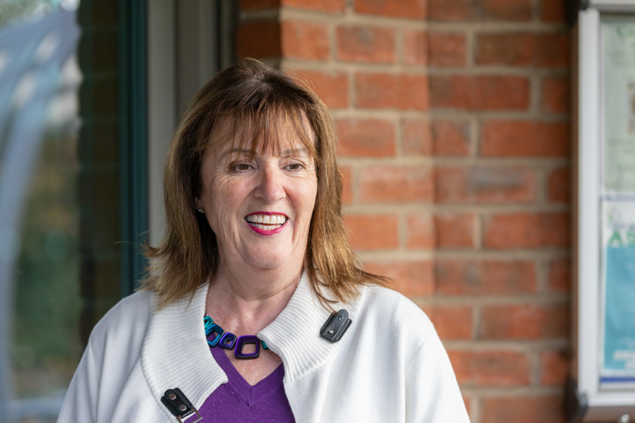 A female looks away from the camera and has a smile on her face. She wears a purple jumper and white coat.