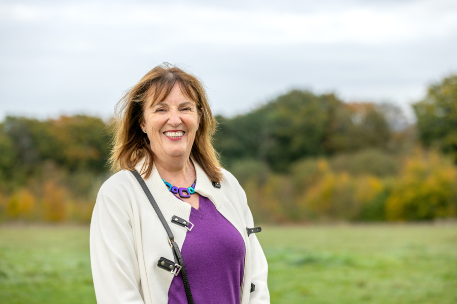 A female looks towards the camera but stands on an angle. She wears a purple jumper and white coat. She is smiling. In the background are trees and grass.