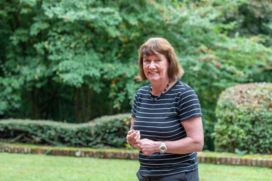 A female looks towards the camera but stands at an angle. She wears and black and white striped T Shirt and has a green garden area behind.