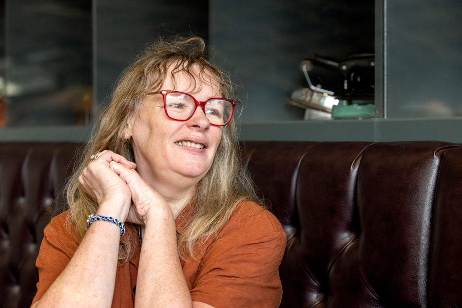A femle wears red glasses and an orange top. She is sitting down looking to the right with her hands clasped up by her face.