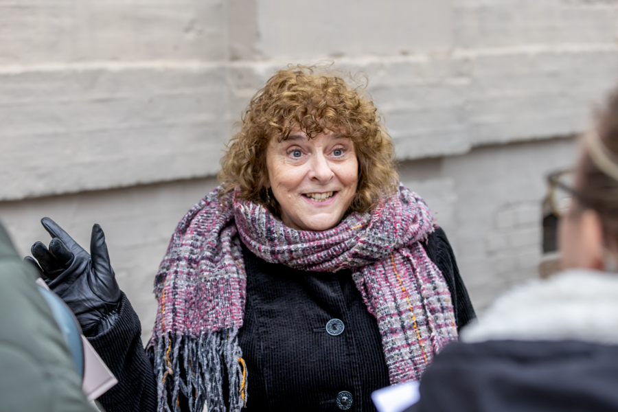 A females wears a black coat and gloves and a brightly coloured scar and stands talking to others.