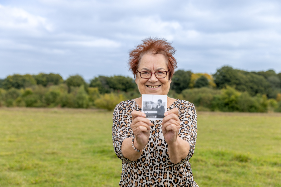 A female stands wearing a leopard print top in the park. She holds out a photo of her younger self.