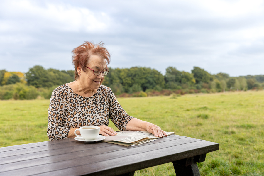 A female sits wearing a leopard print top in the park. She has a cup of coffee next to her and looks through a scrapbook.