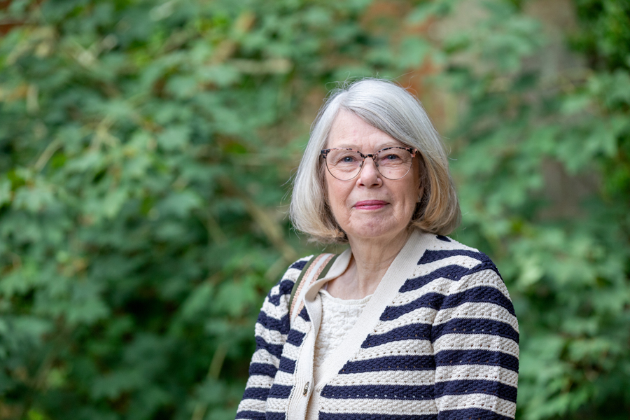 A female stands wearing a black and white striped cardigan. There is greenery in the background..