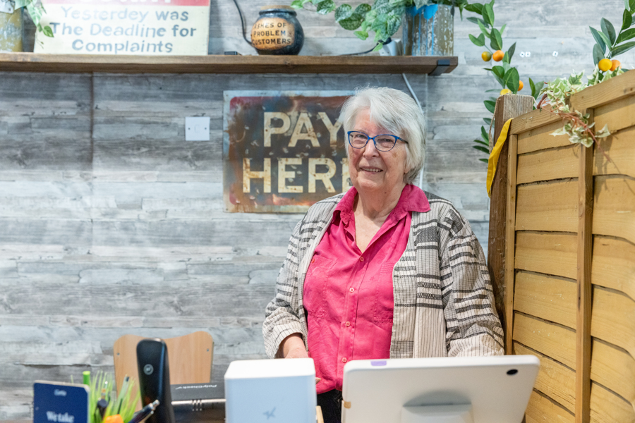 A female wearing a bright pink shirt and grey and white patterned cardigan. She is in a shop standing behind the till.