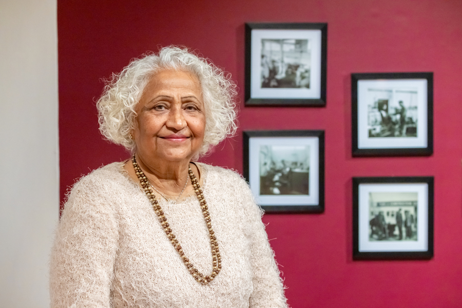A female stands in front of a red wall which has photos on it. She is smiling and looking at the camera.
