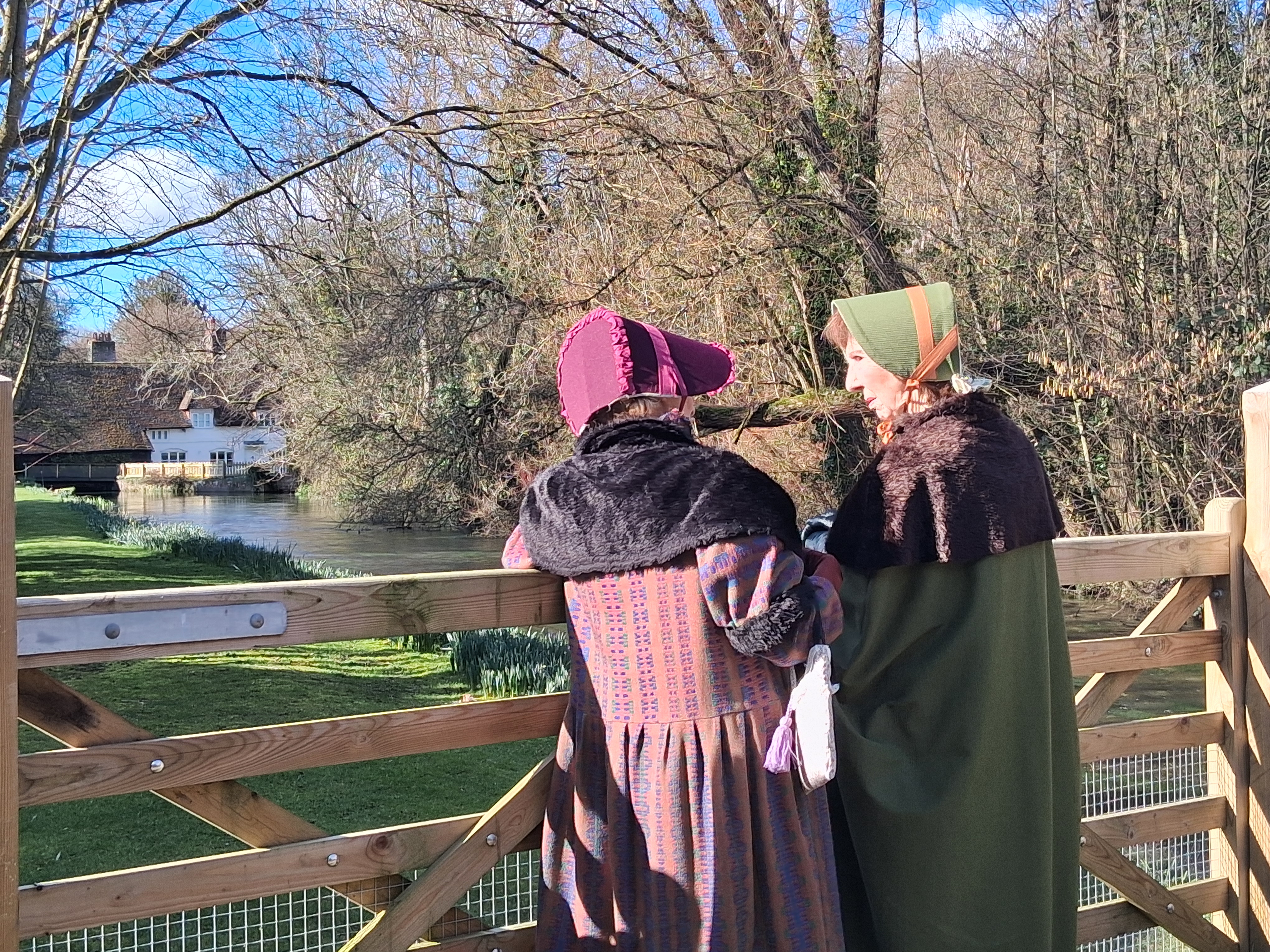 Two women dressed in regency clothing are on the Overton trail. You can see the river and an old building in the background.