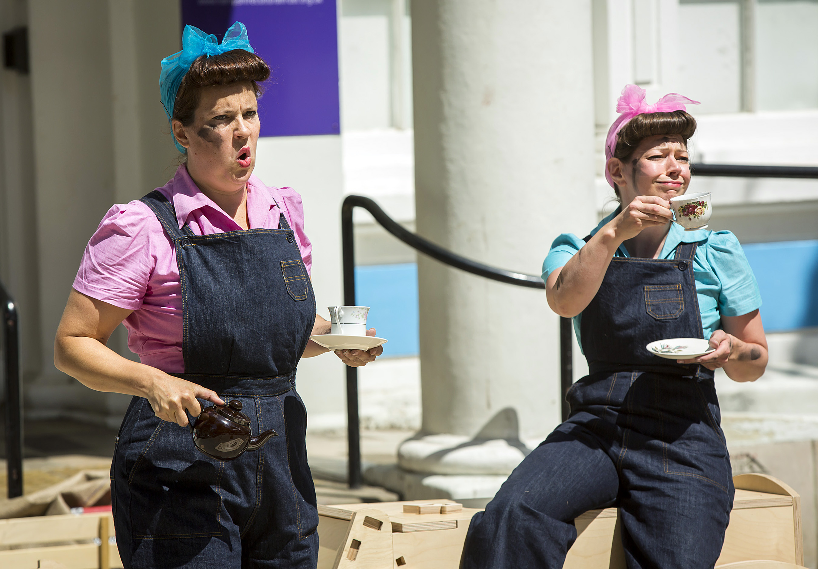 Two females dressed in dungarees with their hair tied back in bows hold cups of tea.