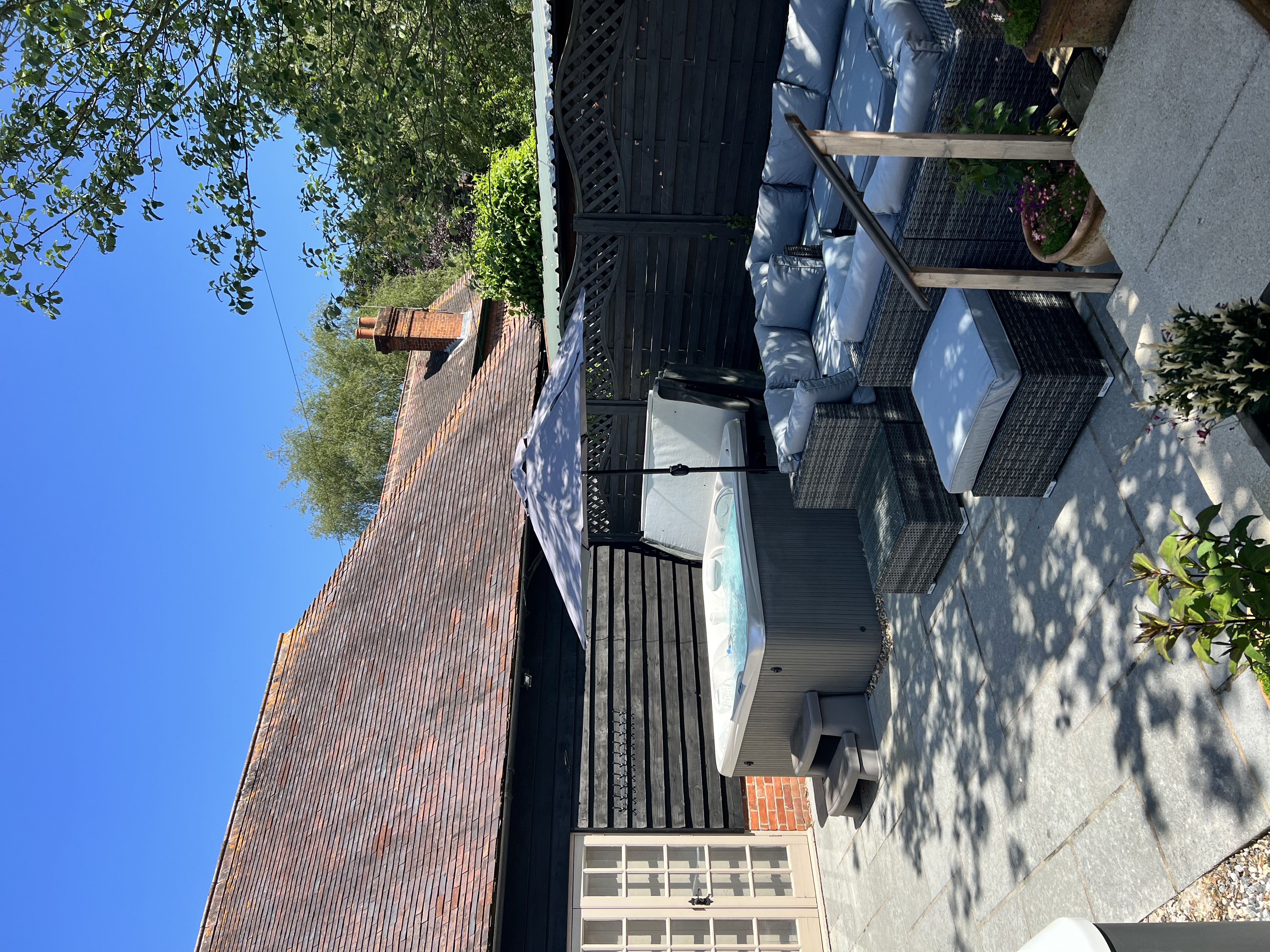 An external image of day spa Laurabella in Basingstoke. The image shows a seating area and hot tub on a paved patio. The sky is blue and in the backdrop is the rear of the building, a tiled roof with wooden cladded walls.