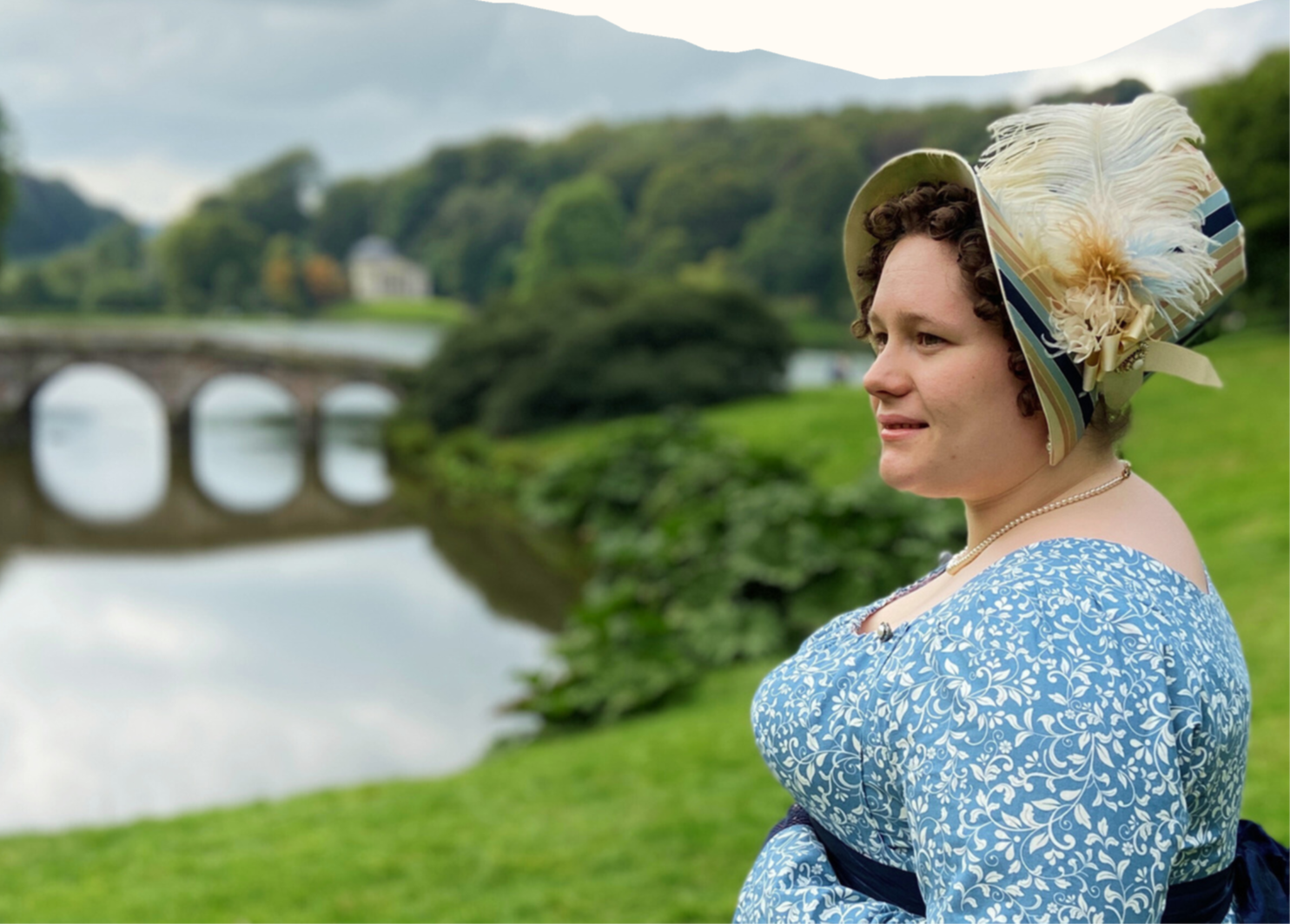 A lady is stood on a grassy riverbank  overlooking a river She is wearing a white bonnet and a blue dress..