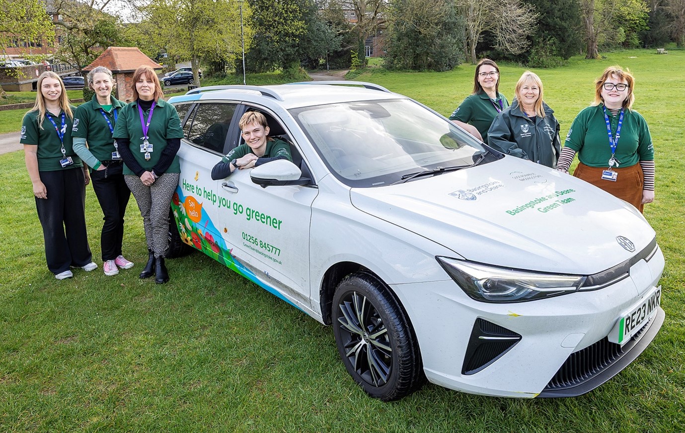 Six people wearing green T shirts stand around an electric vehicle. One person sits inside leaning out of the window.