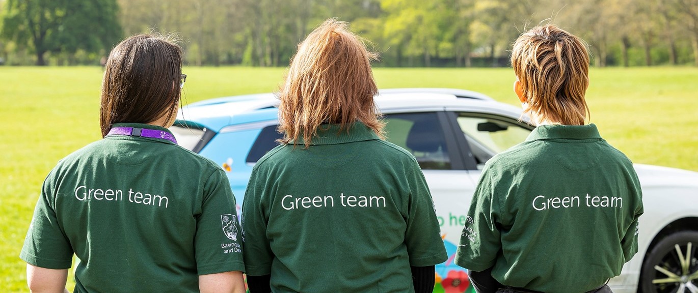 Three people wearing green T Shirts that say Green team on the back stand facing away from the camera with their vehicle in the background.