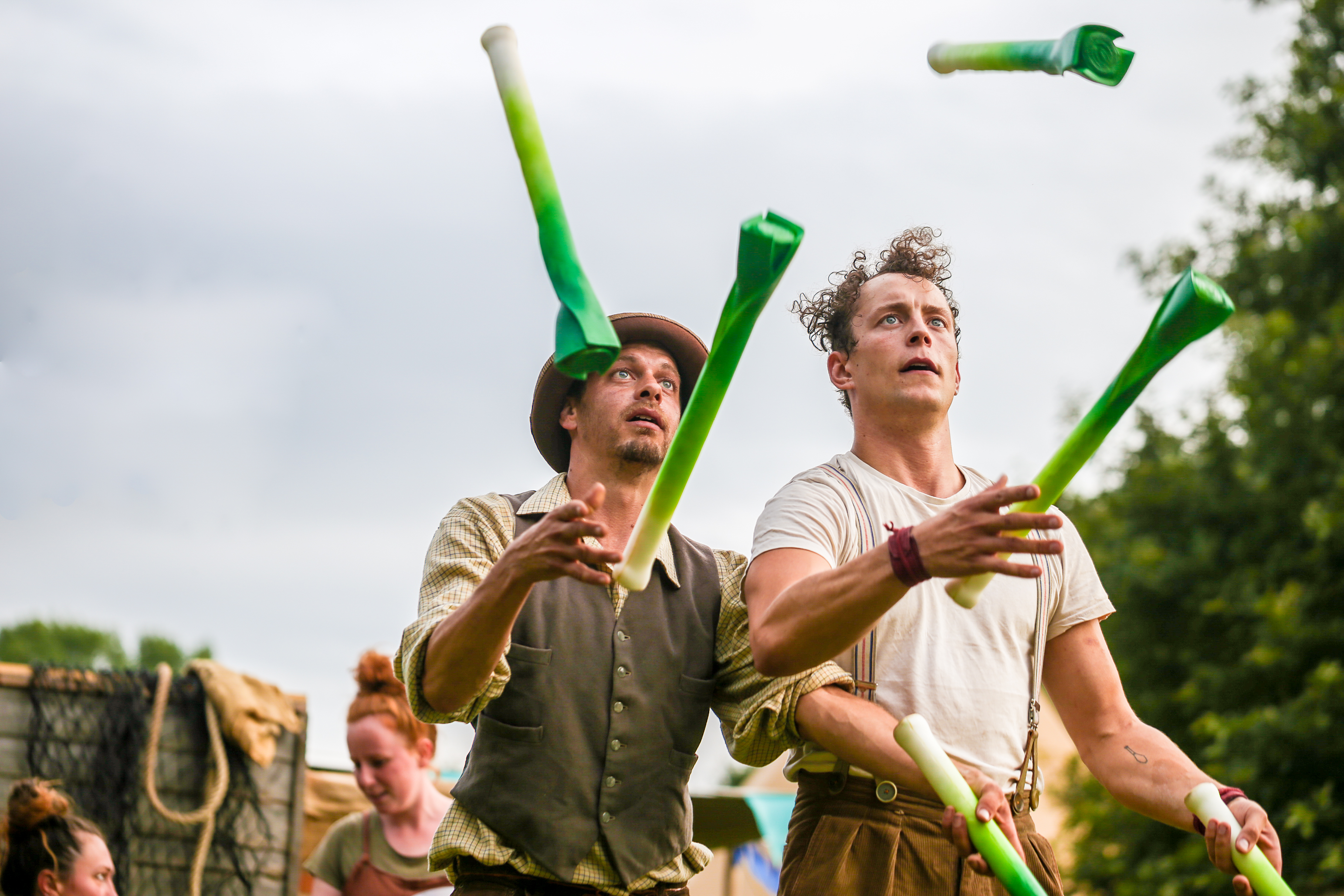 Two performers juggle leeks (the vegetables) They wear brown and cream clothing and both have a concentrated look on their face.
