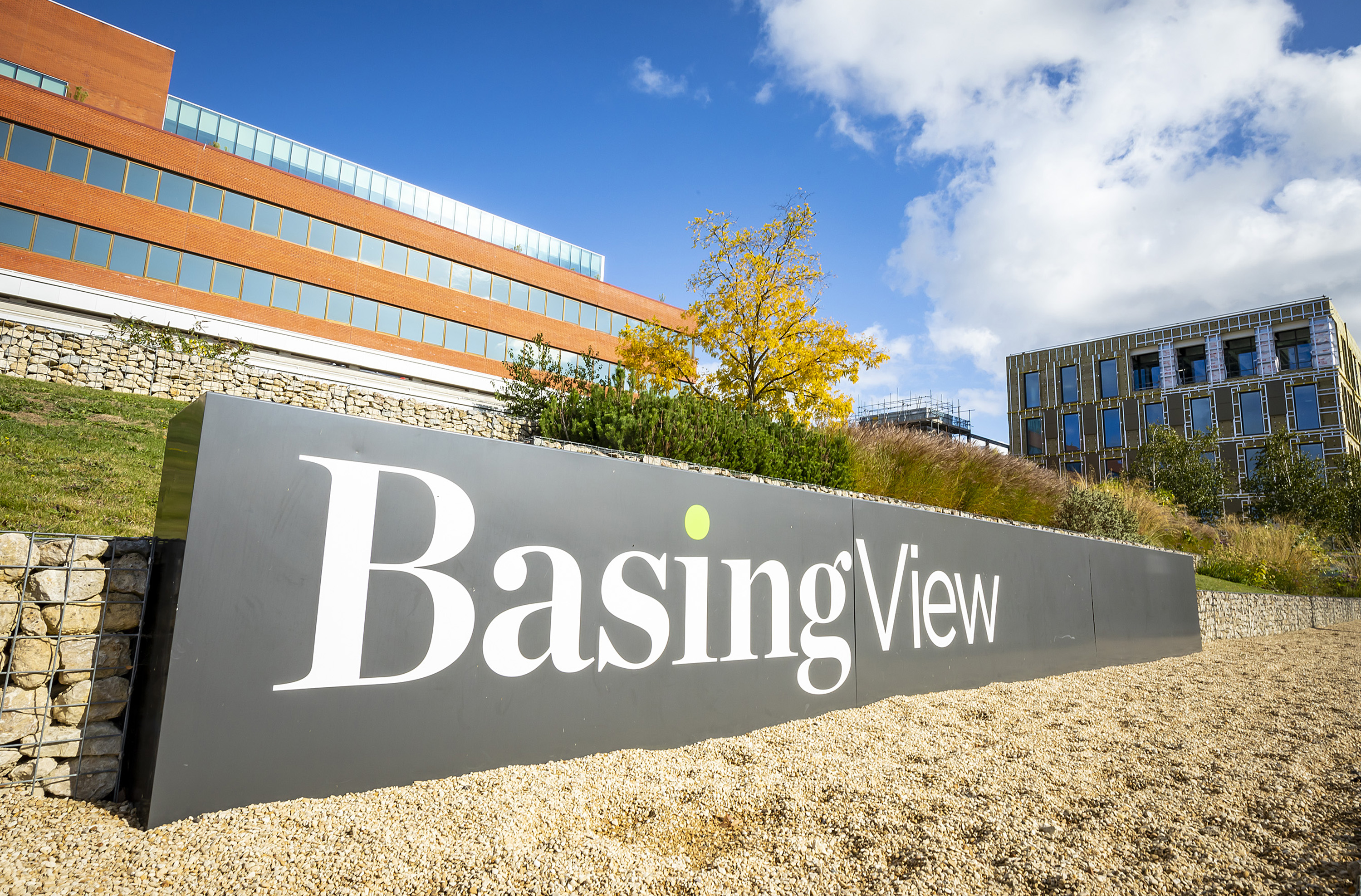 A large Basing View sign, white lettering on a black background. With a red building in the left background and a part built building to the far right in the background.