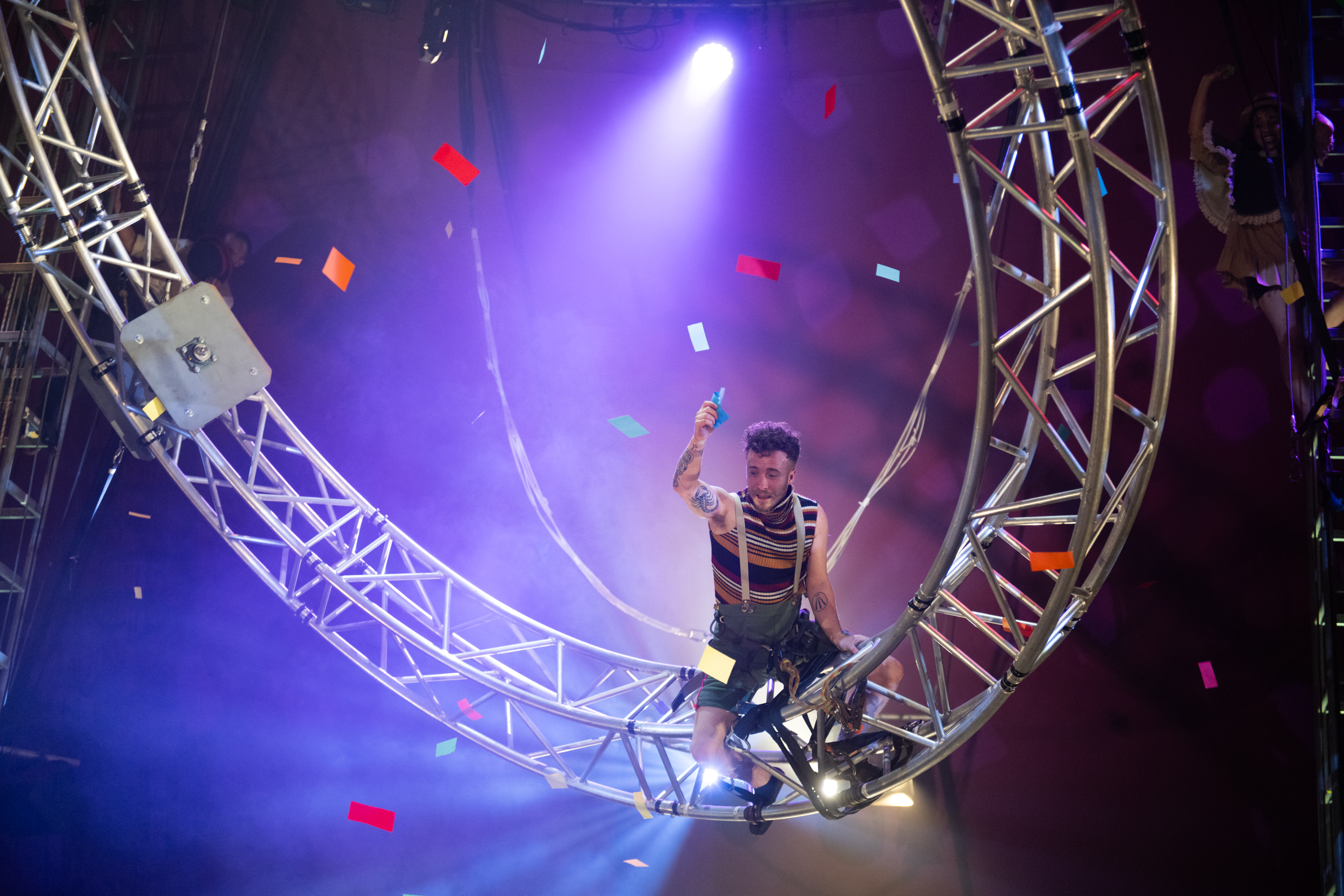 A circus perfomer completes an aerial performance within a dark Big Top Tent. There are speckled lights in the background.