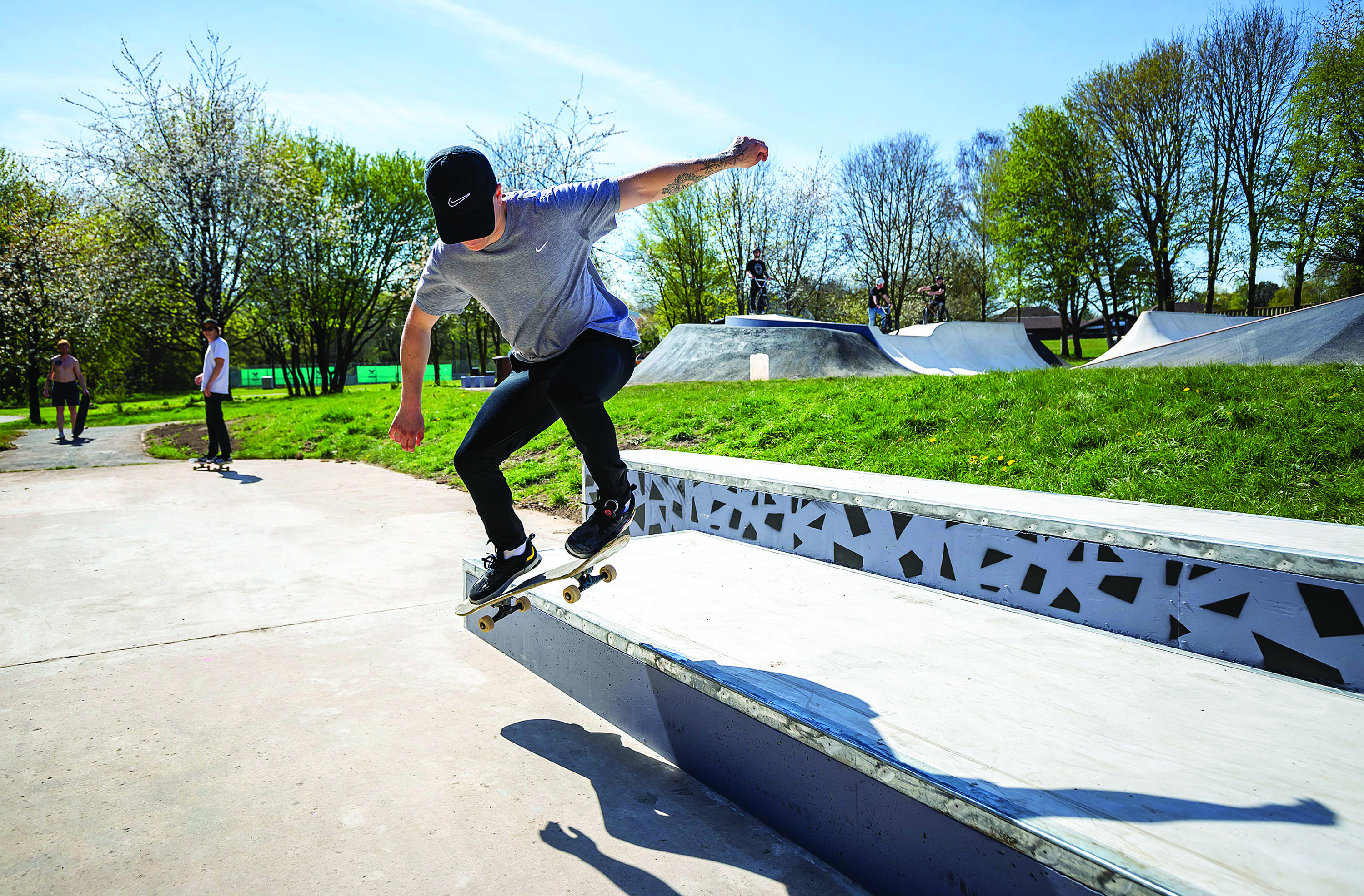 A skateboarder wears black jeans, a grey t shirt and black cap. The skateboarder is midway through a trick at a skatepark. There is grey concrete to the lower half of the photo and blue sky and green grass to the top half of the image. Two skateboarders in the distance look on to the camera.