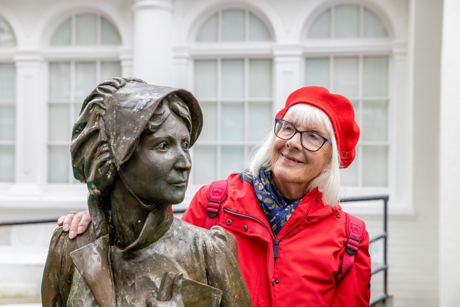 A woman in a red coat and a red hat is smiling and is stood behind a statue of Jane Austin.