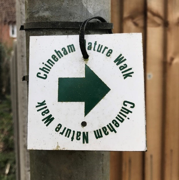 A close up photograph of a white square sign with a green arrow pointing right to the Chineham Nature Walk. The sign is clipped to a metal post. The background is blurred.
