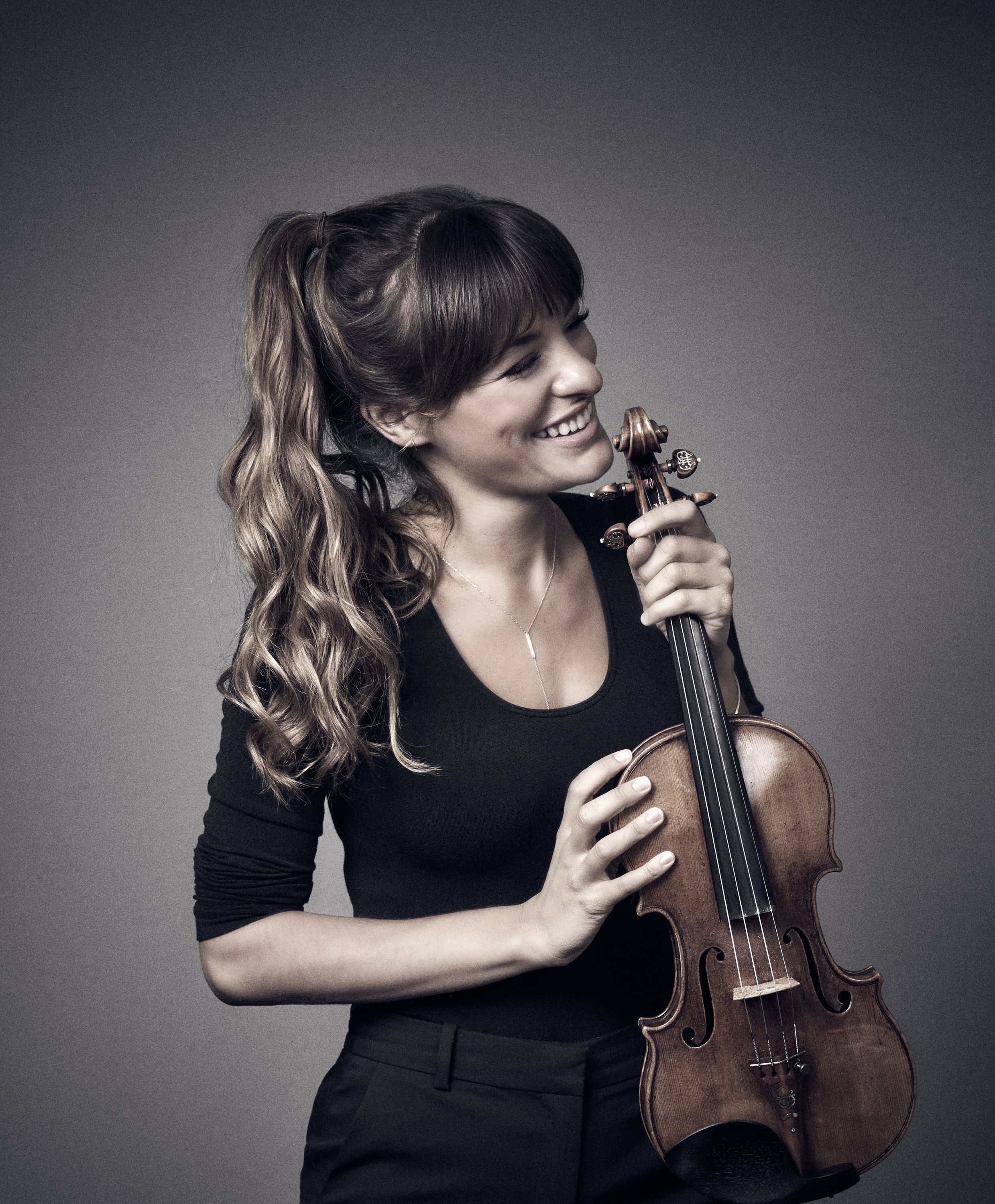 A lady in a black dress with her hair tied back is holding a brown violin