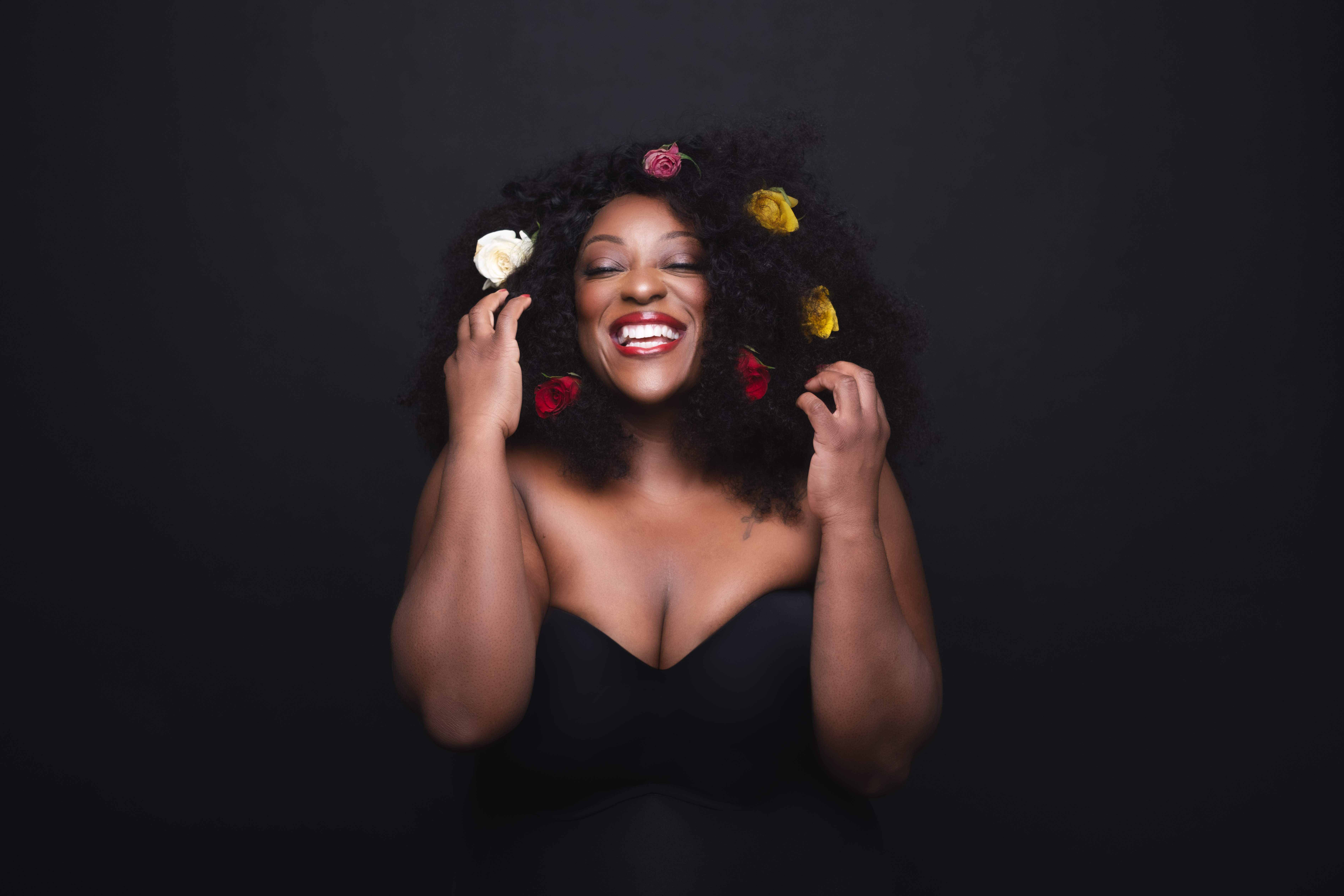 A female wears coloured flowers in her black heair and a black bodice style top. She appears against a black background.