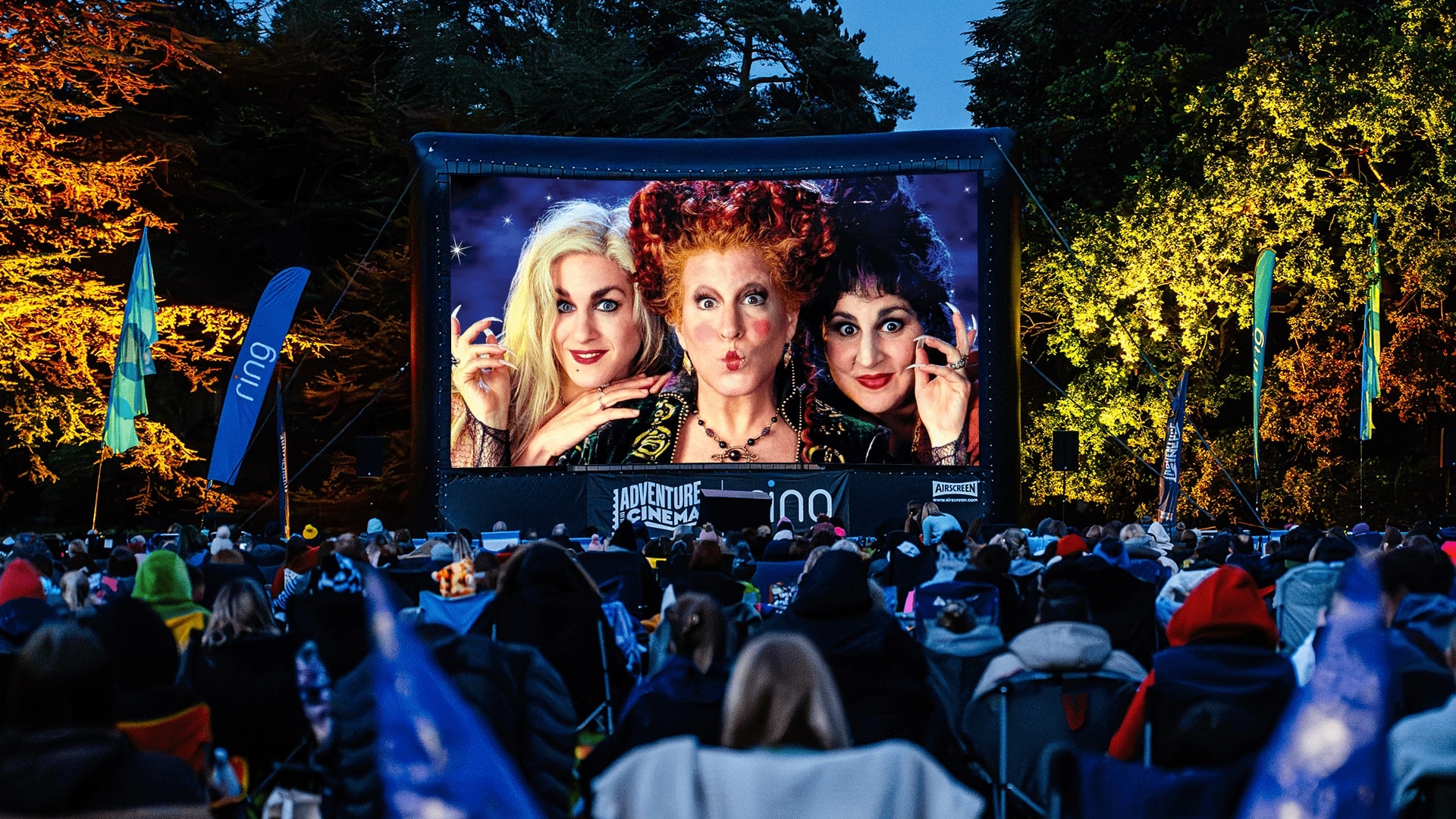 A film screen with three females on. In front sit people on deck chairs. There are lit up trees either side.