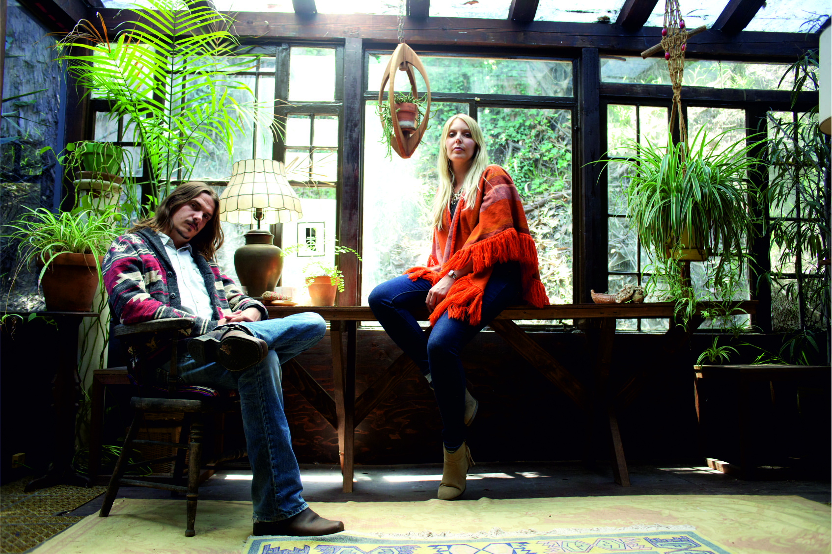 A man with longbrown hair is sat on a chair. A woman in an orange jumper is sat next to the man, sitting on a wooden table.