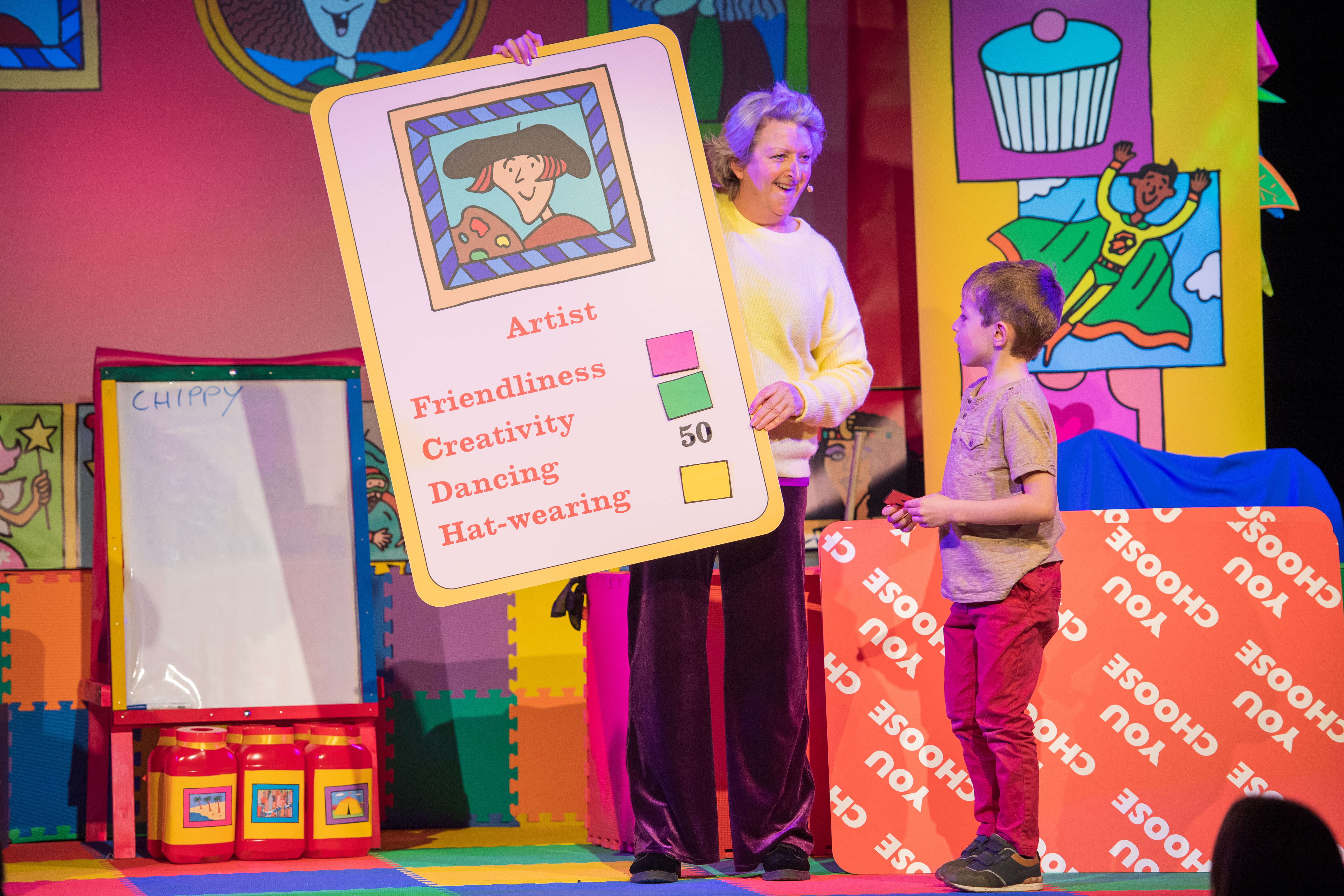 A woman is stood on stage in front of an audience. She is holding a large card and talking to a young boy with short brown hair.