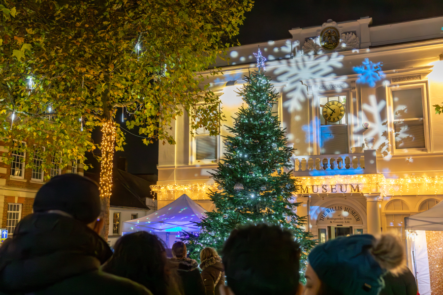 A crowd of people gather at Market Square in Basingsoke town centre. They watch blue and white lights projected on The Willis Museum.