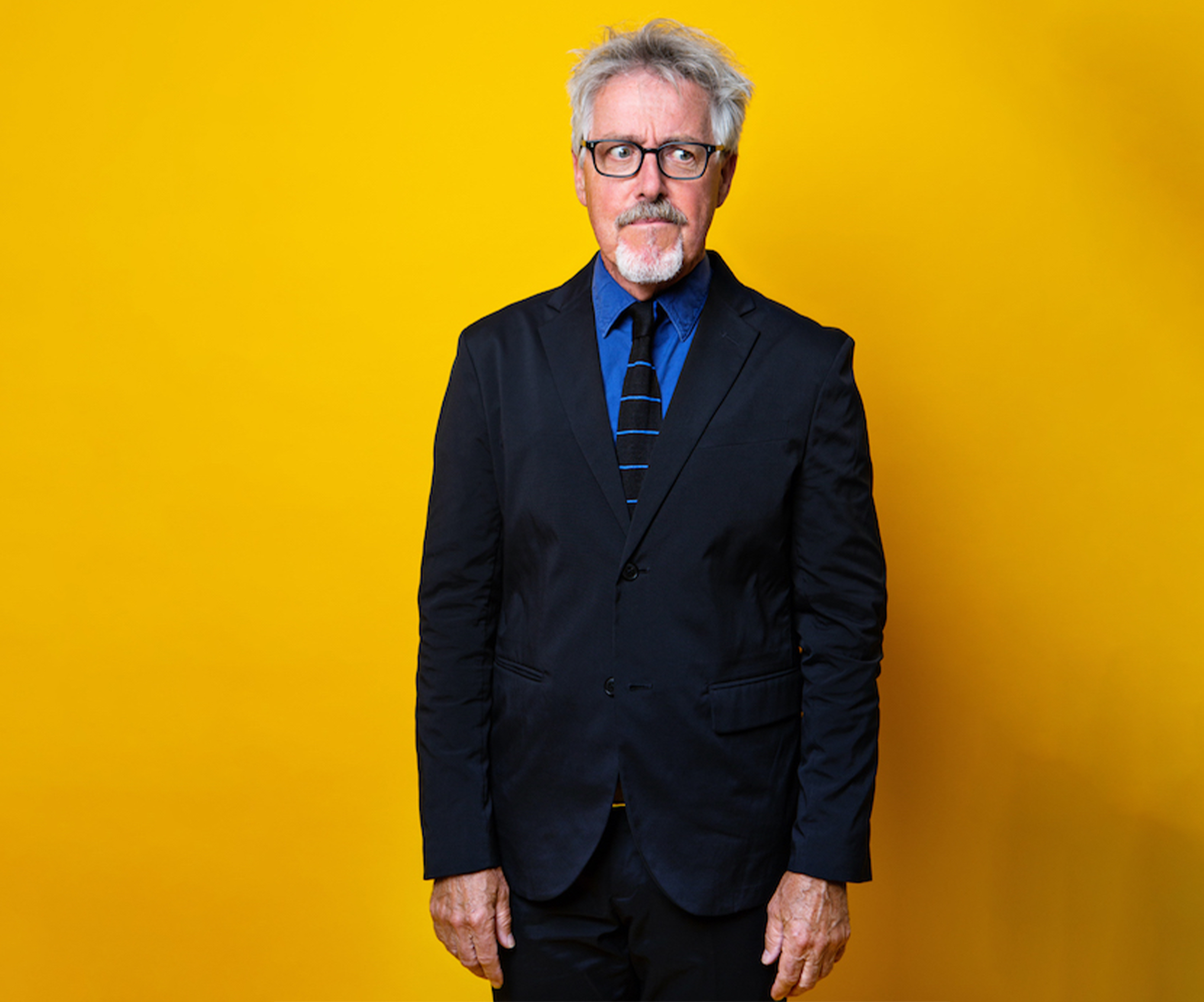 A man with grey hair. He is wearing glasses and a black suit with a blue shirt and a black and blue stripped tie. He is stood in front of a yellow background.
