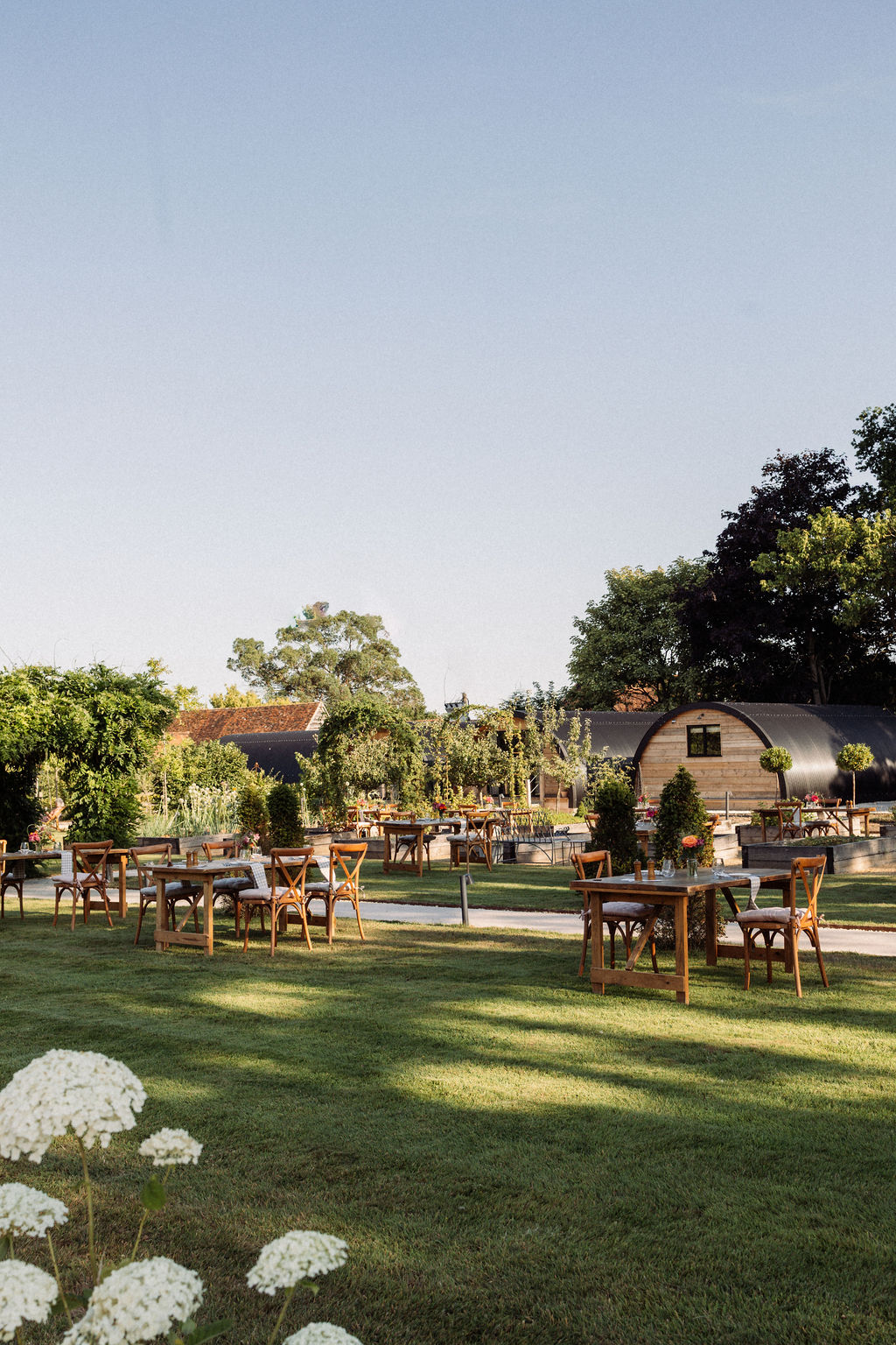 A grassed area with white flowers in the foreground. Behind you can see tables set out on the grass and some accommodation behind.