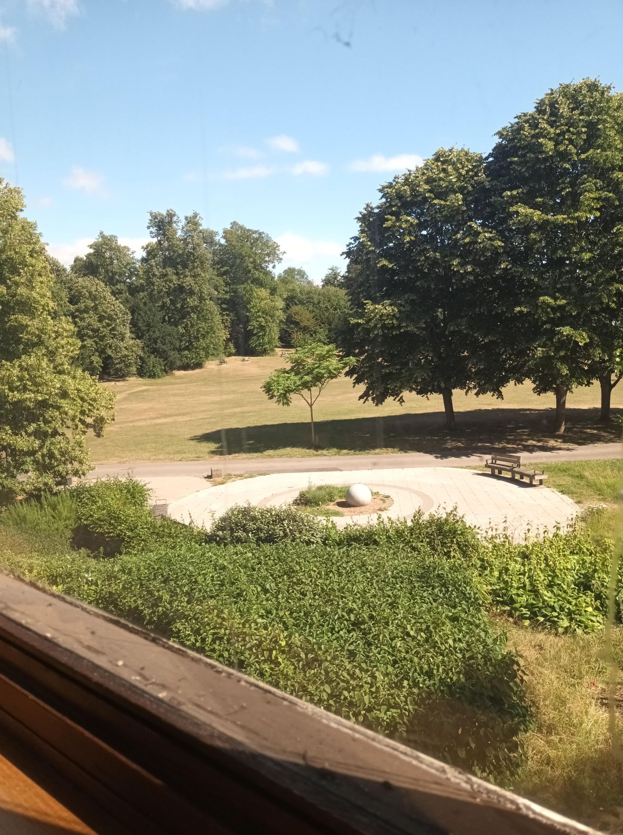 The wooden window frame. You can see a round paved area with a round stone in the middle and some benches. Beyond this are grass and some very tall trees.