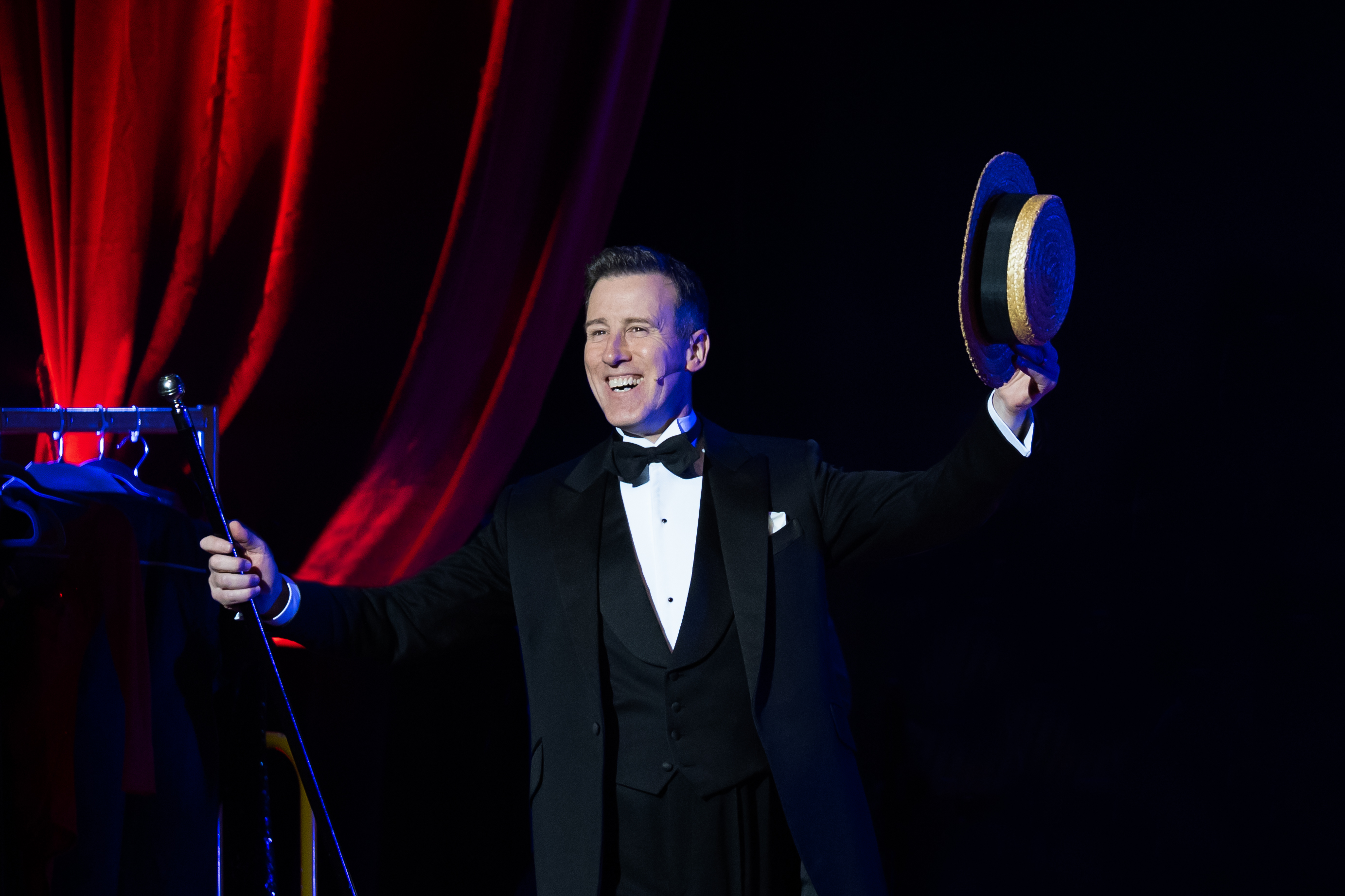 A man is dressed in black tie and holds a cane and hat. In the background is a red curtain.