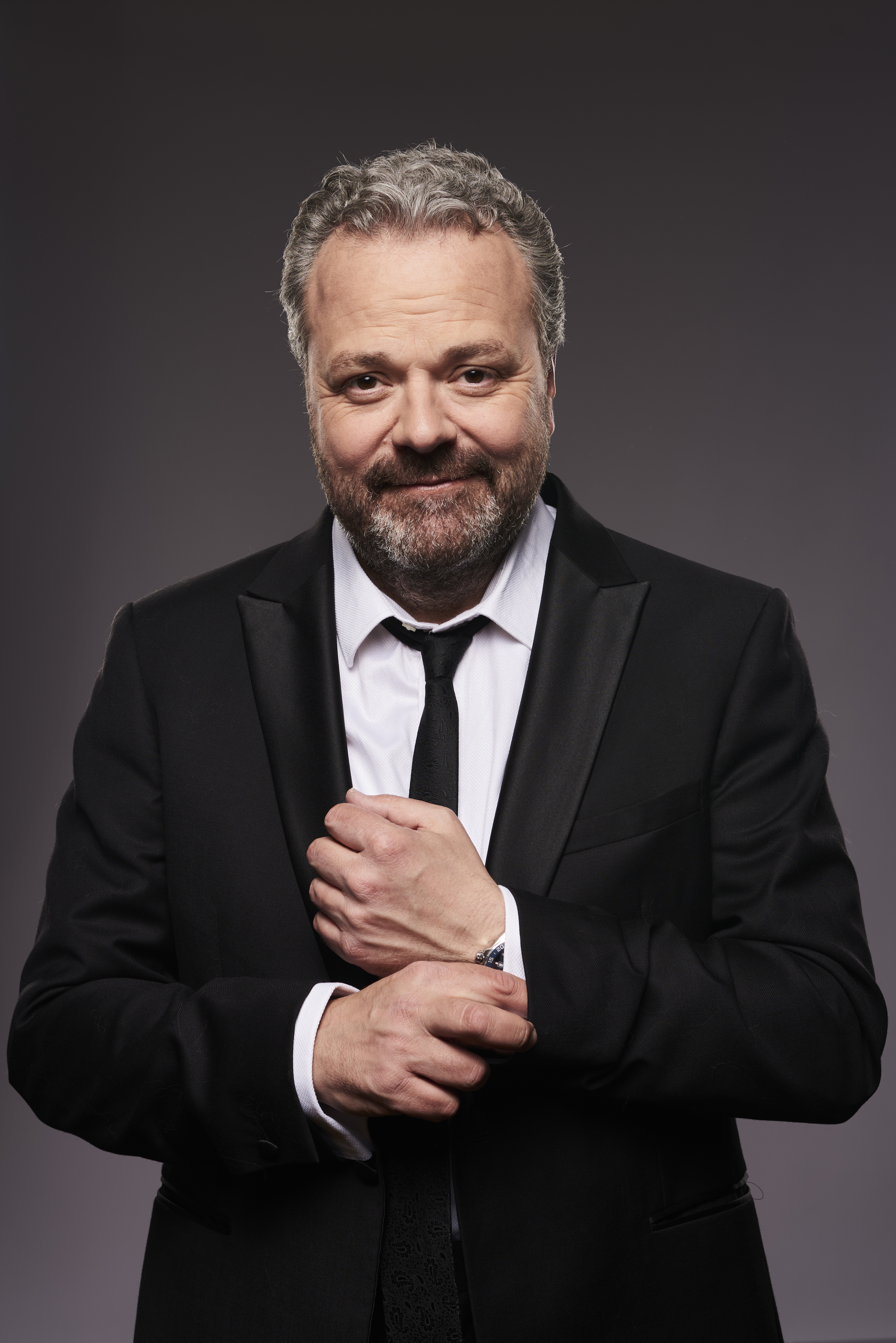 A man with short grey hair. He is stood in front of a grey background wearing a balck suit and a black tie. He is smiling at the camera.