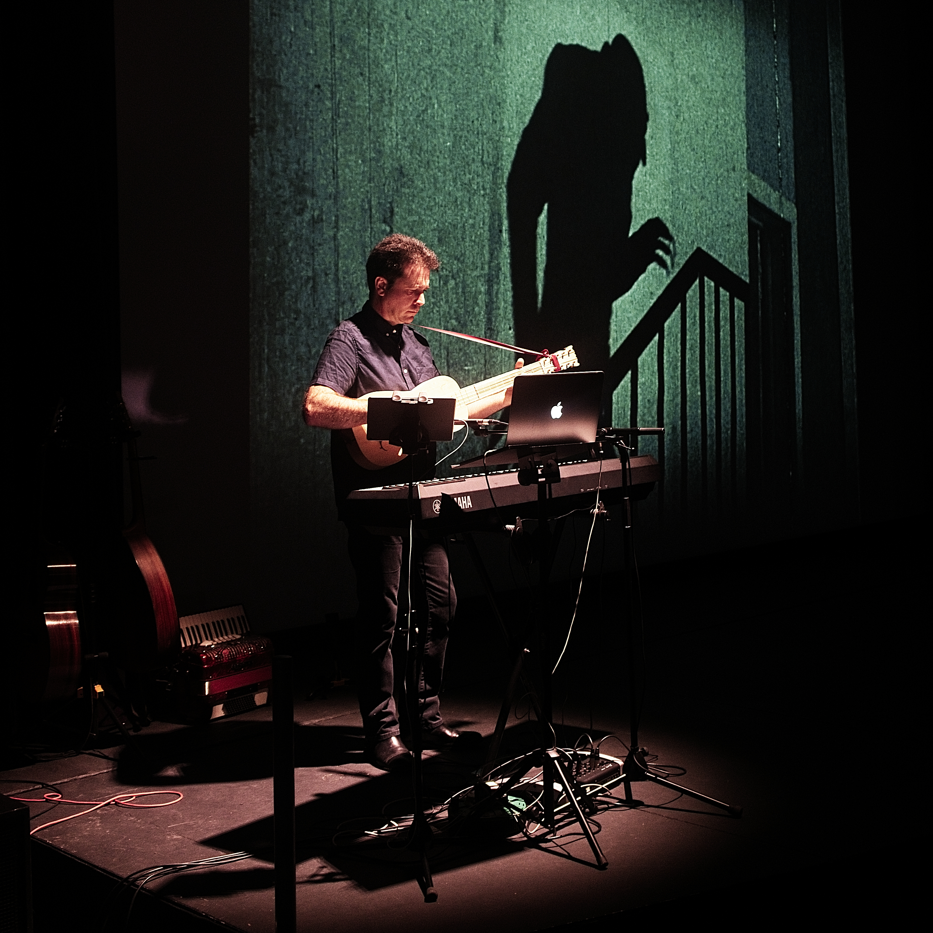 A man is stood up in a darkly lit room. He is playing the guitar and stood behind a keyboard on stage.