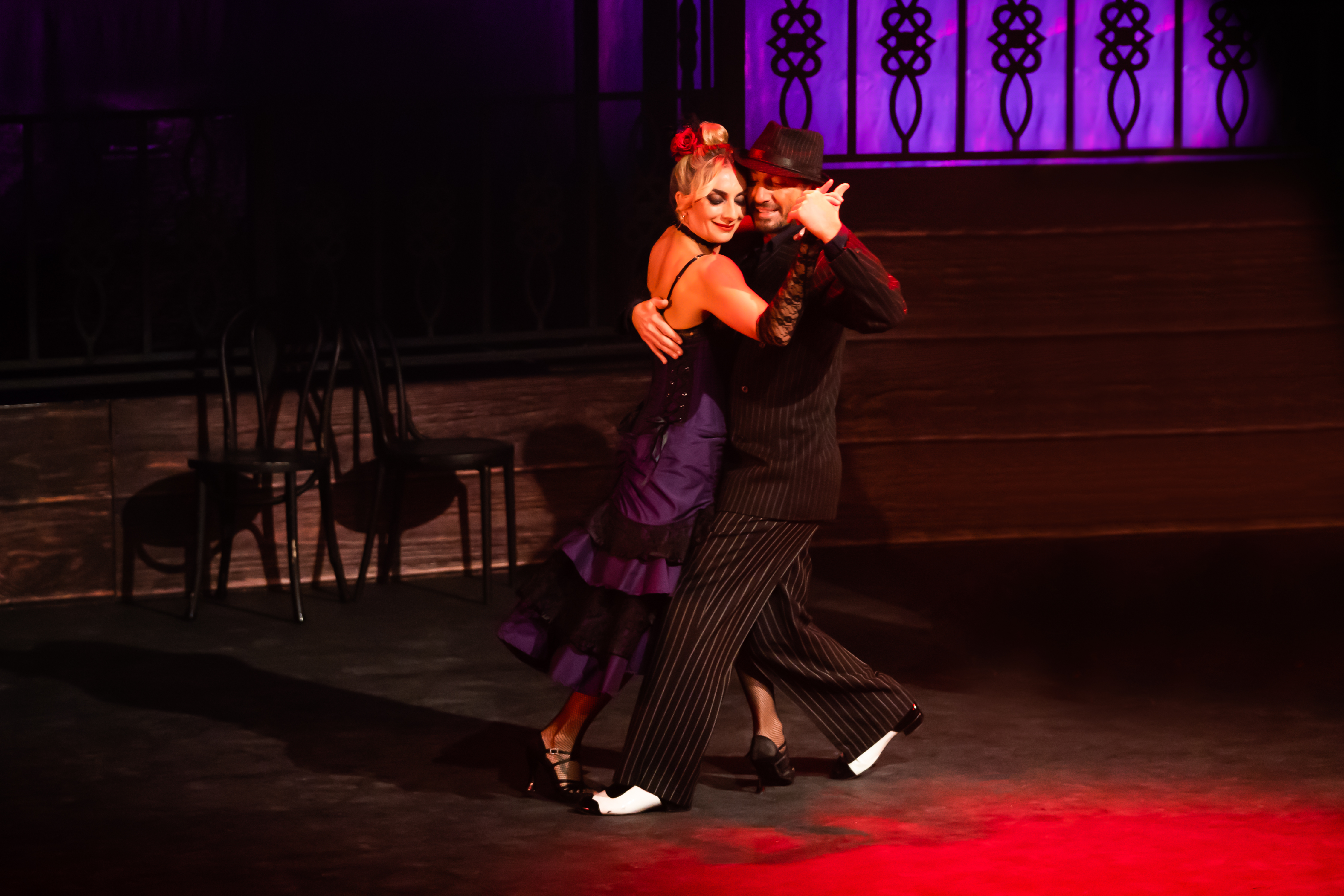 Argentine Tango King and Strictly Come Dancing superstar, Vincent Simone is on stage dancing the Tango with a back with short black hair in a black siuit. She is wearing a dark purple dress and her hair is in a high bun.