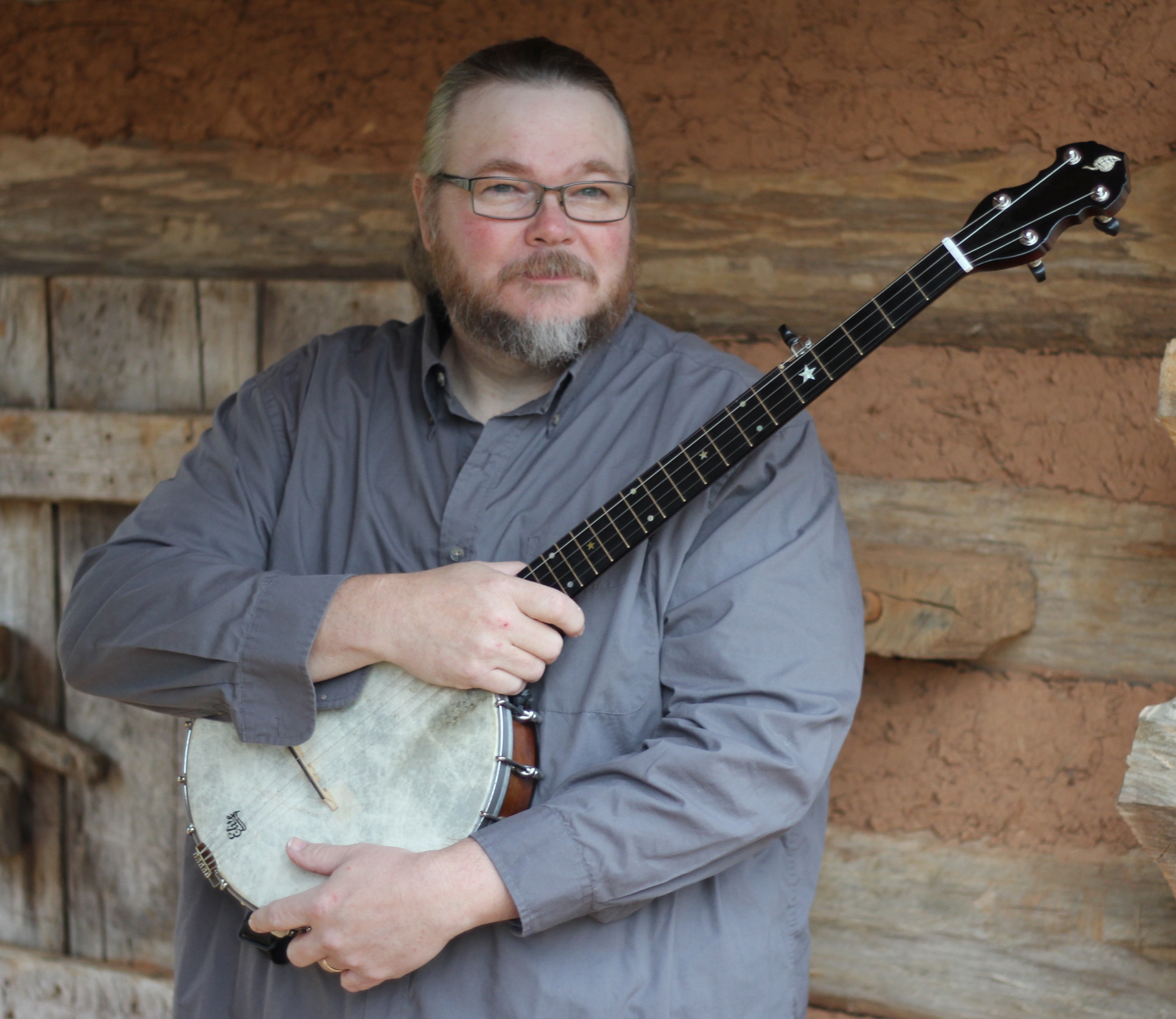 A man who has grey hair. He is smiling at the camera and he is wearing a light grey shirt. He is holding a ukelele that is brown and white in colour. He is stood in front of a wooden door.
