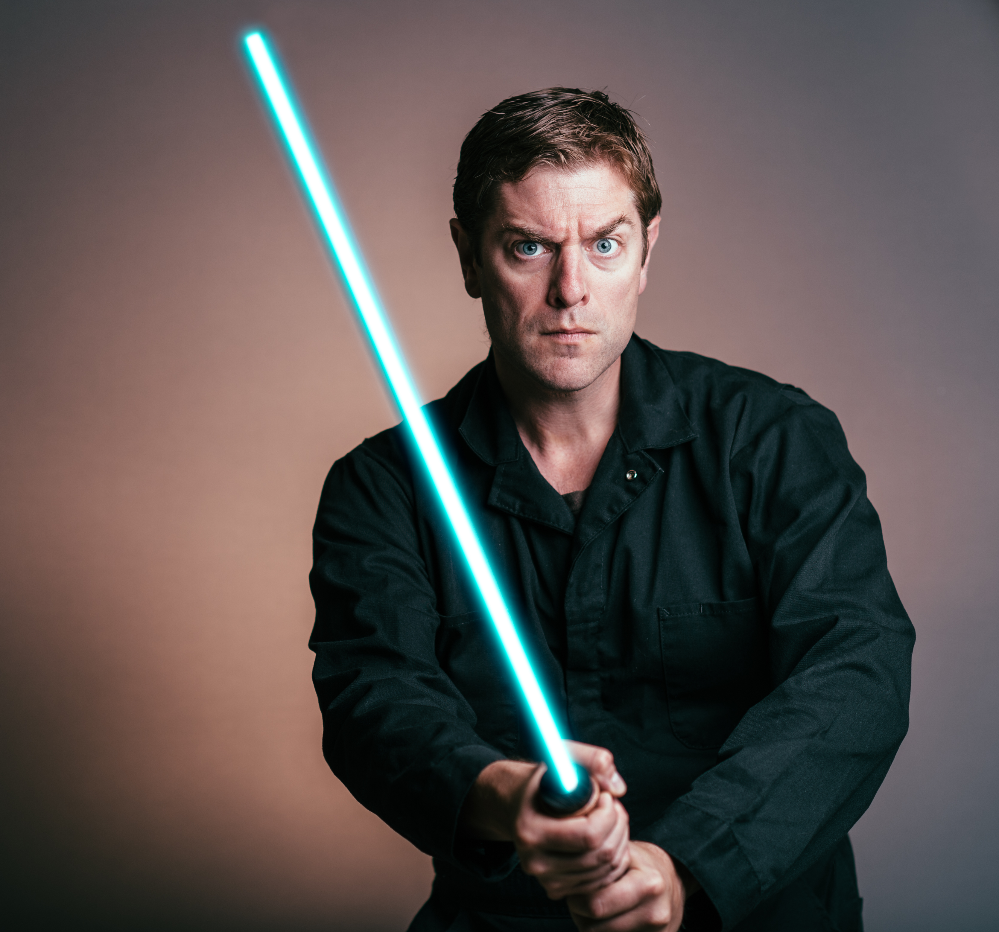 A man with short balck hair is looking at the camera with a serious face expression. He is wearing a black shirt and he is holding a lit up blue light saber.