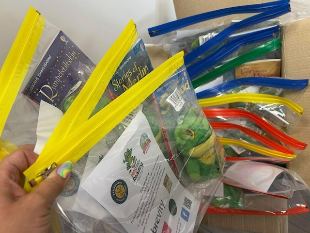 A hand holds some clear bags that contain a book, piece of paper and cuddly frog.