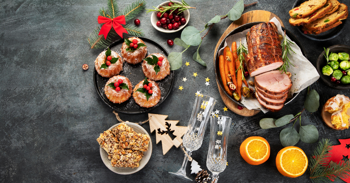 A dark grey table cloth. A plate of festive mince pies. A large gammon. Two orange halfves. A bowl of red canberries. A wooden Christmas tree.