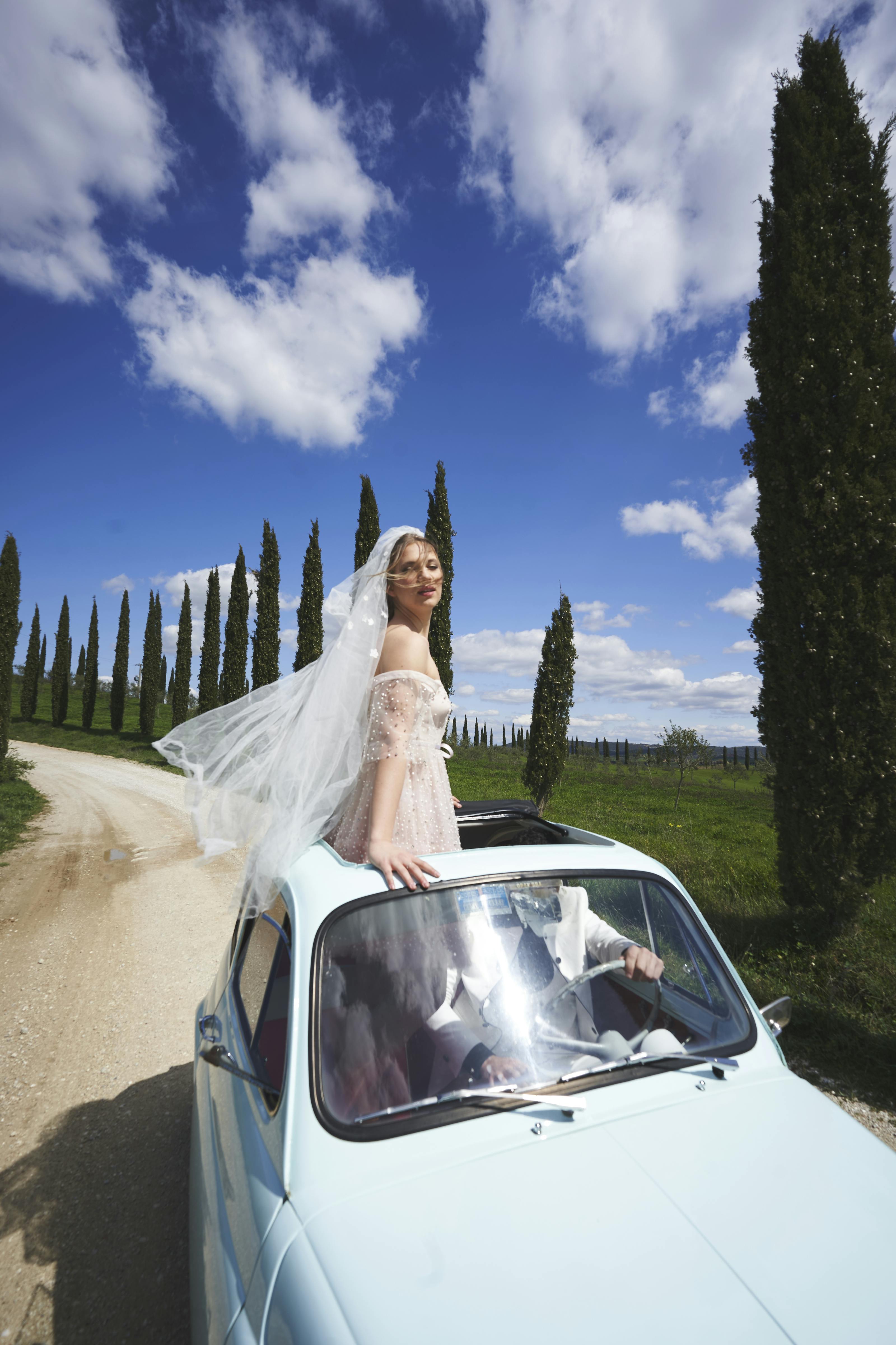 bride on the roof of vintage fiat 500 on a countryside road with blue sky