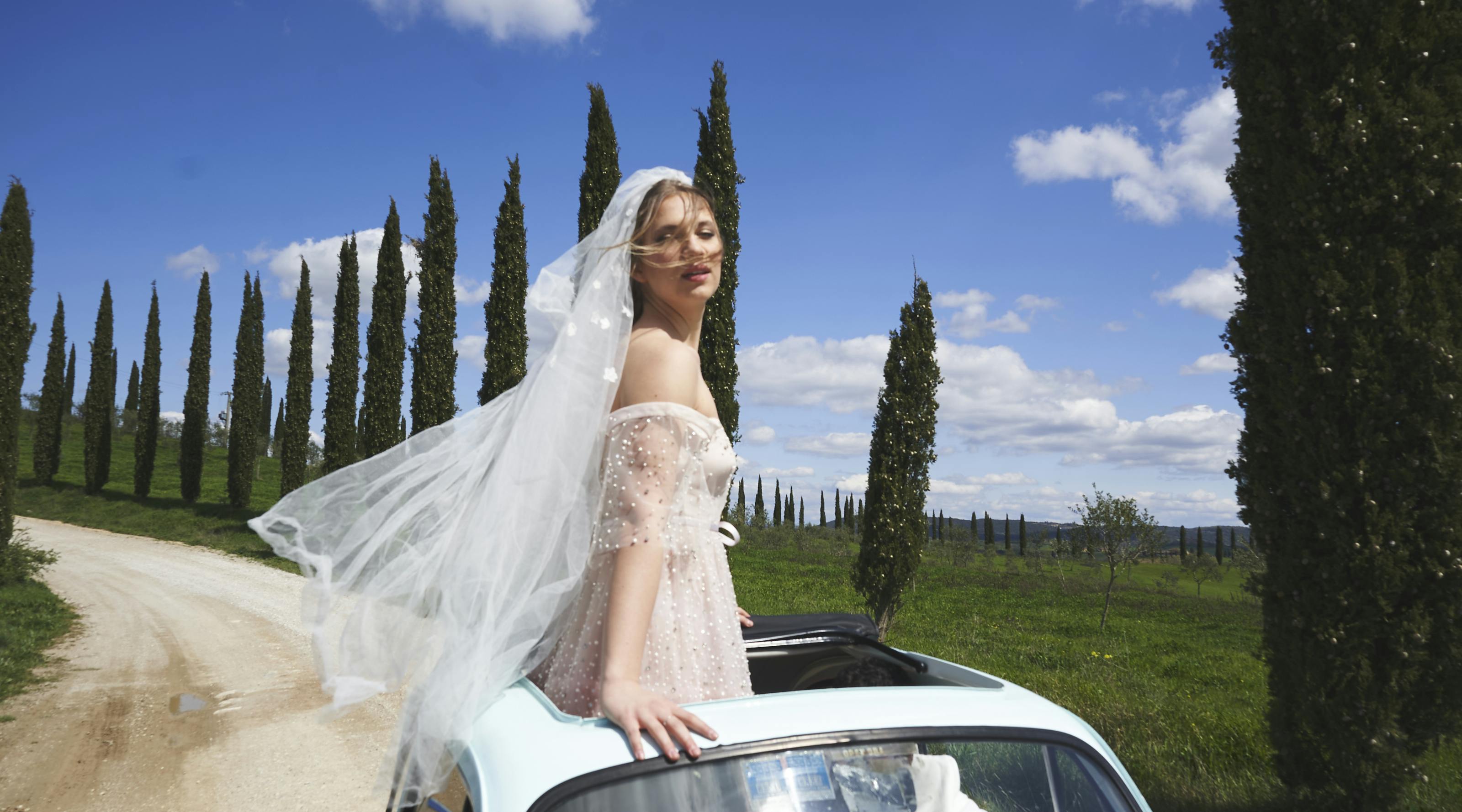 bride on the roof of vintage fiat 500 on a countryside road with blue sky