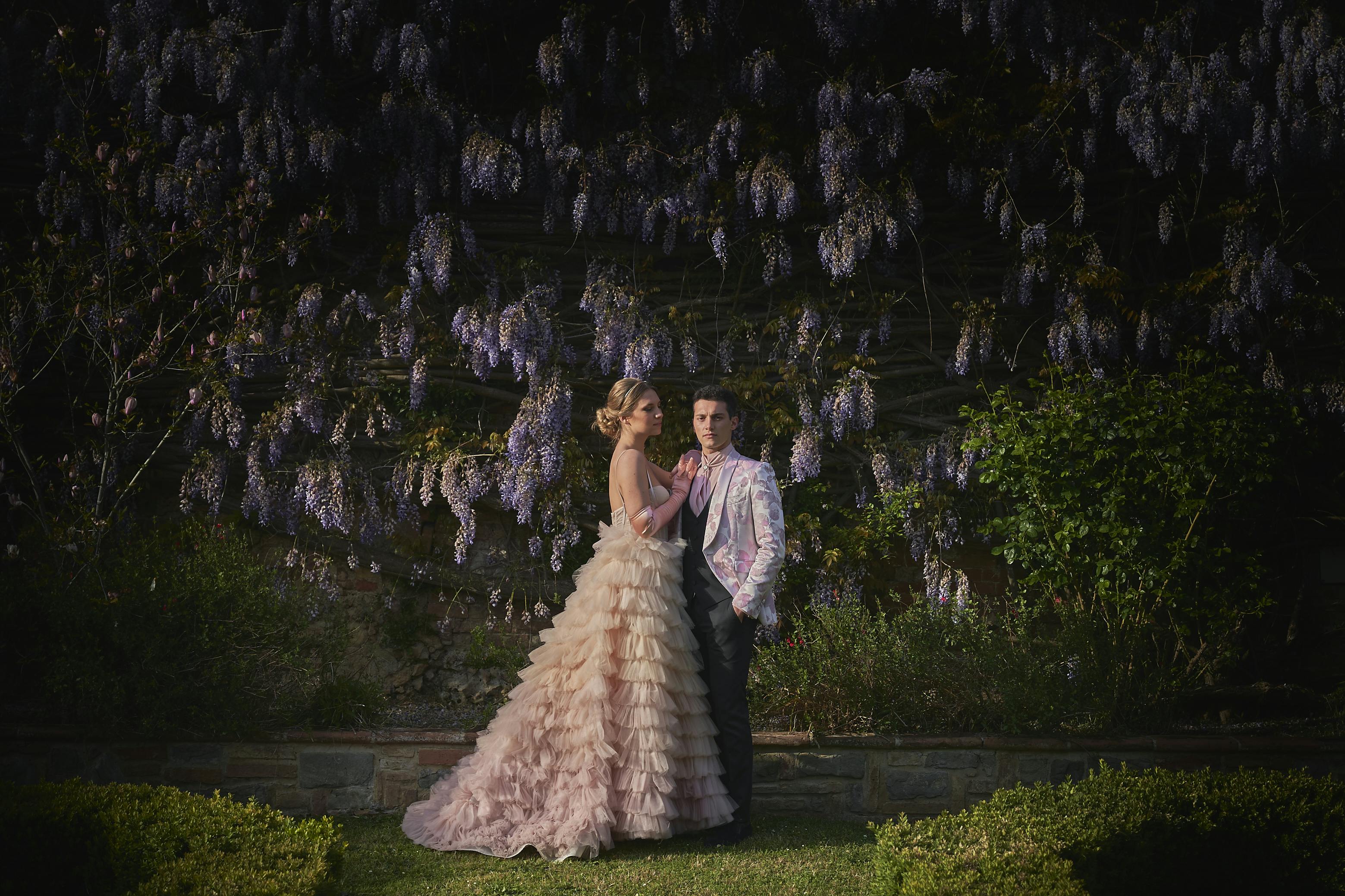 bride and groom dressed in fashion wedding dresses in front of a wall of wisteria plantsll of
