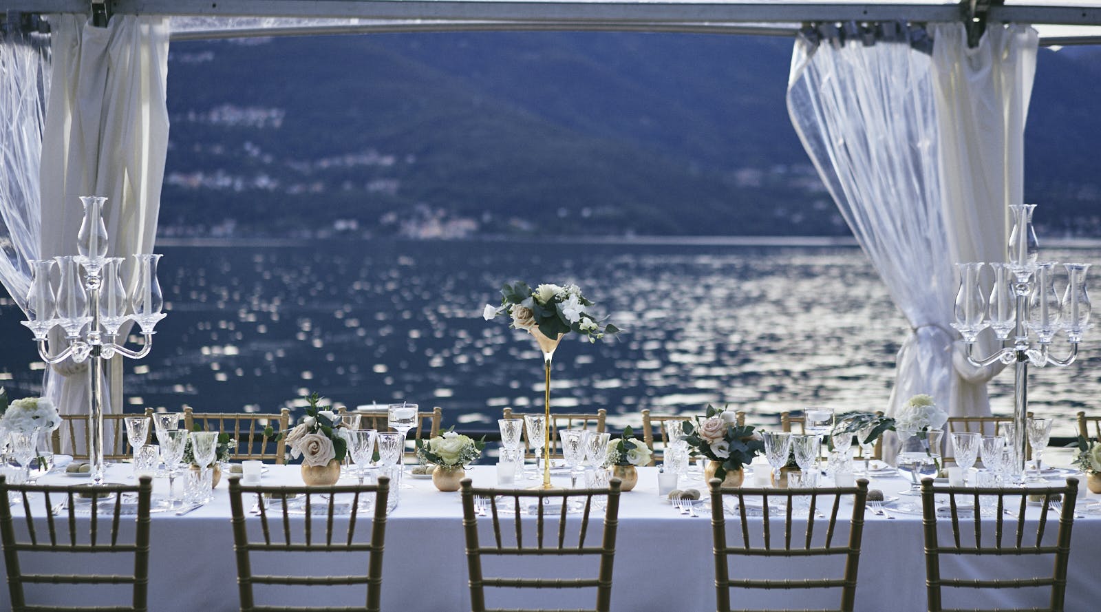 Tavola apparecchiata per matrimonio di fronte al lago con sedie oro e bianche centrotavola tende bianche