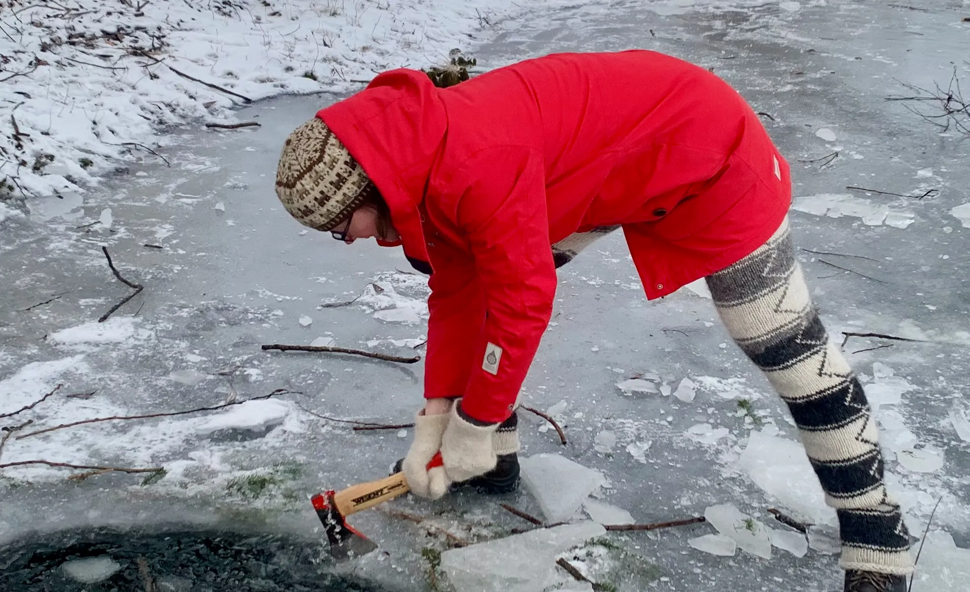 A woman in a red coat, knitted wool pants, and a knitted wool hat uses a hatchet to break the ice on a frozen lake.